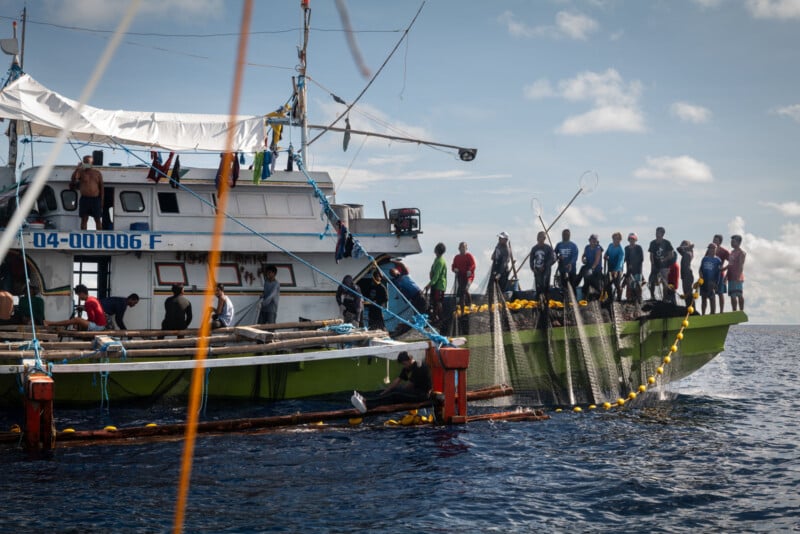 A group of people work together on the deck of a fishing boat, hauling in a large fishing net from the ocean under a partly cloudy sky. Some crew members are pulling the net aboard while others observe.