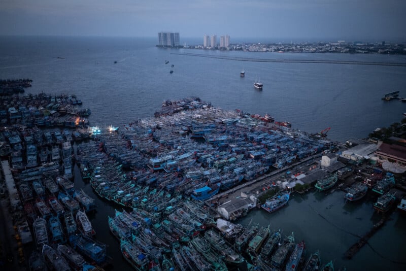 Aerial view of a densely packed harbor filled with fishing boats at dusk, with city buildings and the coastline visible in the background under a cloudy sky.