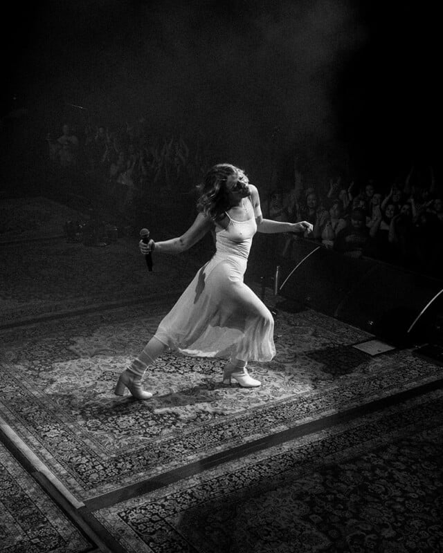Una mujer con un vestido fluido sostiene un micrófono vibrantemente en el escenario y baila en el escenario, iluminada por el centro de atención, y la audiencia anima en el fondo. La imagen es en blanco y negro.