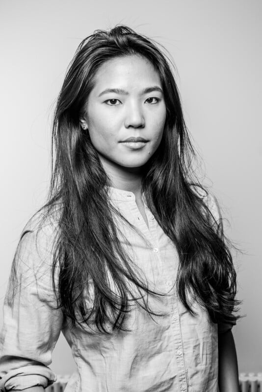 Black and white portrait of a young woman with long hair wearing a light-colored button-up shirt, looking confidently at the camera against a plain background.