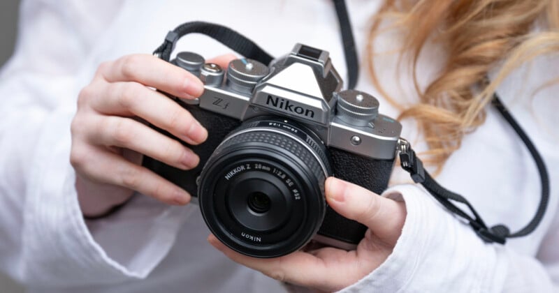 Close-up of a person holding a vintage Nikon film camera with both hands, wearing a white long-sleeve shirt. The camera strap is visible, and the person's wavy hair is partially shown.