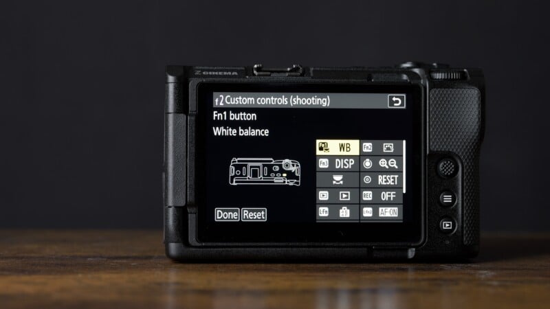 Close-up of a digital camera's rear screen displaying custom controls menu for shooting mode, highlighting the Fn1 button and white balance settings. The camera is on a wooden surface with a dark background.