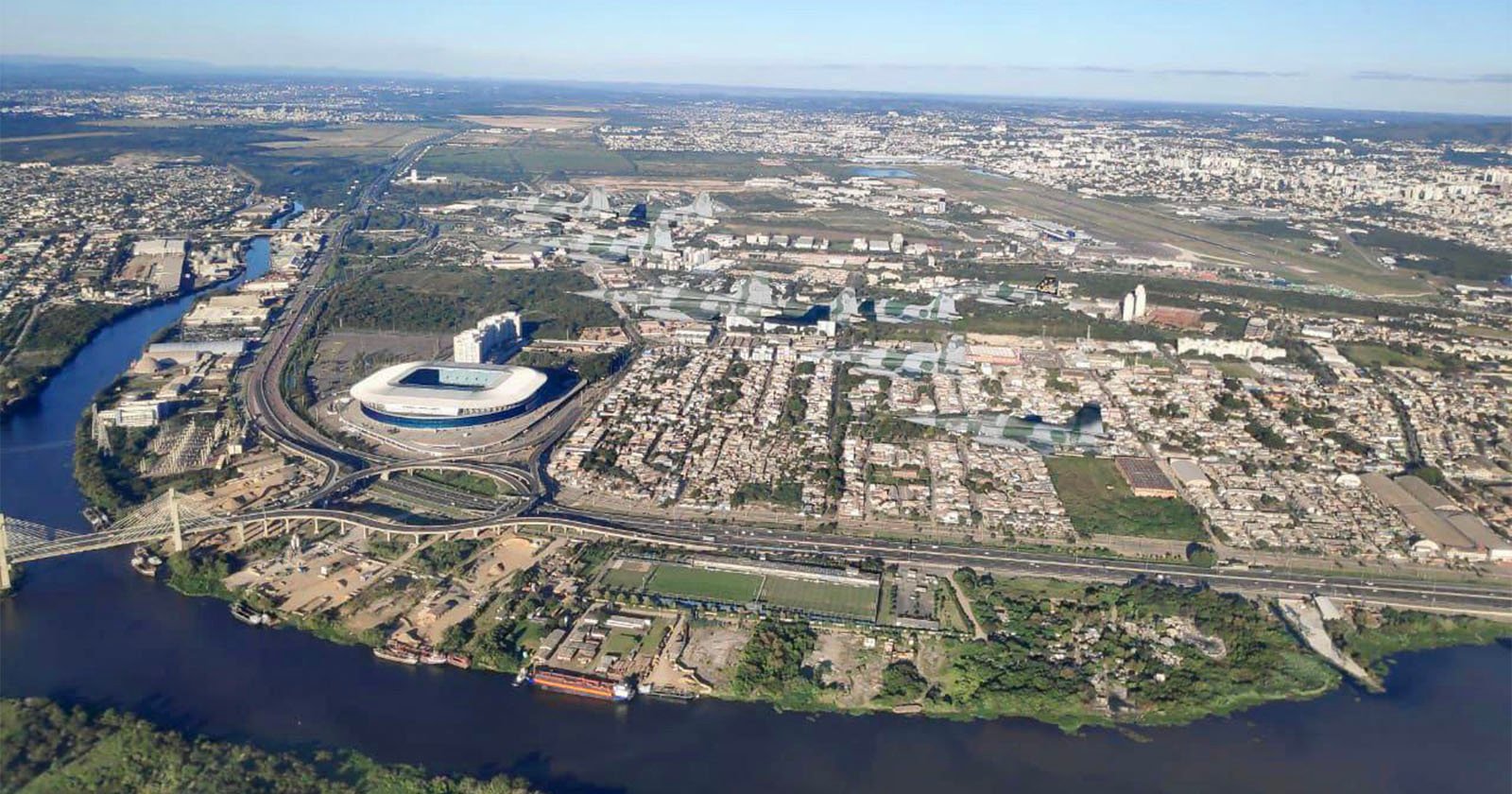 Aerial view of a city with a large stadium near a river, surrounding urban areas, highways, and scattered greenery, with distant buildings and an airport visible in the background.