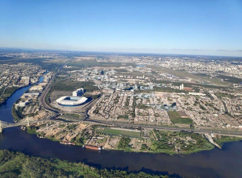 Aerial view of a city with a stadium, river, and urban areas surrounded by greenery and roads under a clear blue sky.