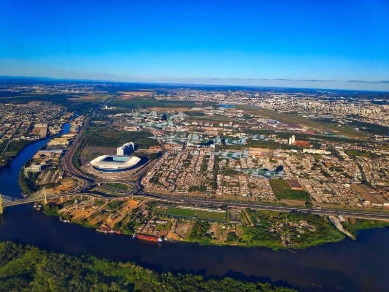 Aerial view of a cityscape featuring a large stadium, winding river, bridge, and dense urban area under a clear blue sky. Green spaces and buildings are visible throughout the landscape.