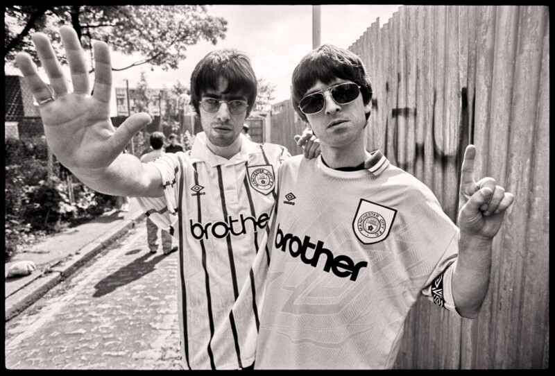 Two men wearing vintage Manchester City football shirts and sunglasses pose on a street; one holds up five fingers, the other holds up one finger. A wooden fence with graffiti and trees is in the background.