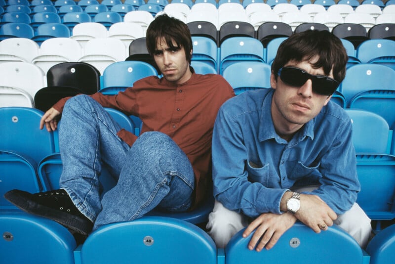 Two men in casual clothes sit on blue and white stadium seats. One wears a red shirt and jeans, the other wears a blue shirt, sunglasses, and a watch. Both look directly at the camera with serious expressions.