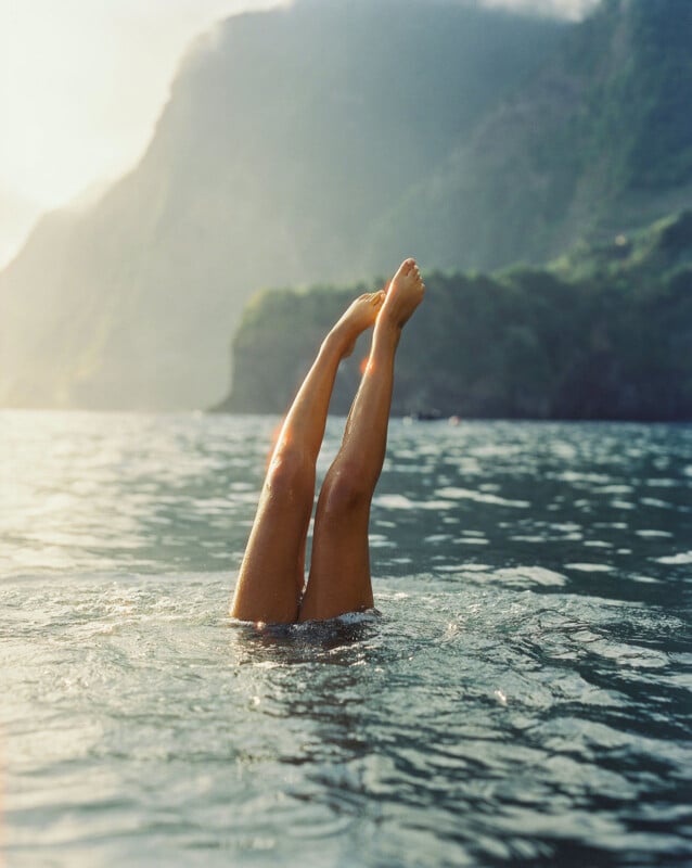 A person is upside down in the water, with only their legs sticking out above the surface, set against a backdrop of mountains and a hazy sky.