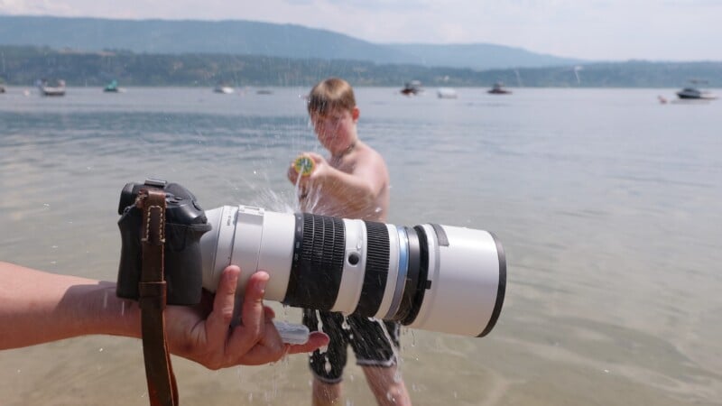 Un niño se paró en aguas poco profundas en el lago, frente a la pistola de agua hacia la cámara grande, arreglando el teleobjetivo en primer plano. Los barcos y las colinas en la distancia se pueden ver debajo del cielo brumoso.
