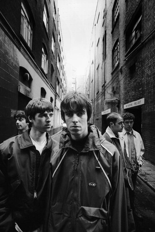 Five young men in casual jackets stand in a narrow, brick-walled alleyway, looking serious. The alley is urban with high buildings on both sides and an overcast sky above.