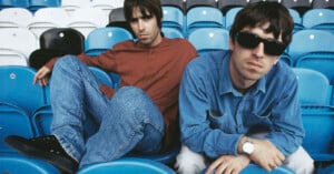 Two men sit among blue and white stadium seats. The man on the left wears a red shirt and jeans with black shoes, while the man on the right, in a blue shirt and sunglasses, leans forward with arms resting on his legs.