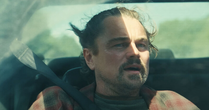 A man with a goatee and tied-back hair sits in a car, looking out the window with a serious expression. Sunlight casts a shadow across his face, and he wears a seatbelt and a plaid shirt.