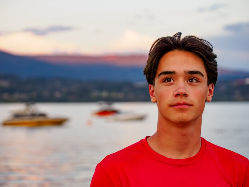 Un adolescente con una camisa roja se encuentra junto a un lago tranquilo al atardecer con botes borrosos y montañas en el fondo.