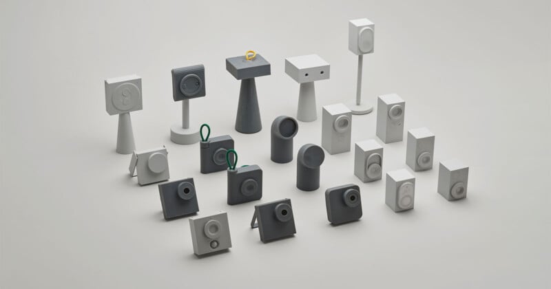 A group of small, abstract geometric sculptures in various shapes and shades of white and gray are arranged on a plain surface. One sculpture has a yellow ring and another has green scissors on top.