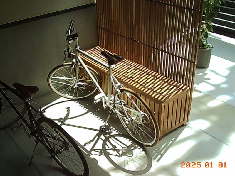 A white bicycle is parked next to a wooden bench in a sunlit indoor area with shadows on the floor. Another bike is partially visible on the left. The date "2025 01 01" is shown in the bottom right corner.