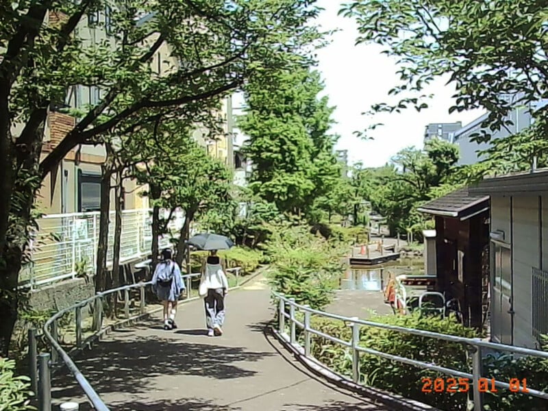 Two people walk along a sunny, tree-lined path beside a narrow canal, with buildings and greenery on both sides. The scene looks peaceful and bright, and the date on the image reads "2023 01 01" in orange.