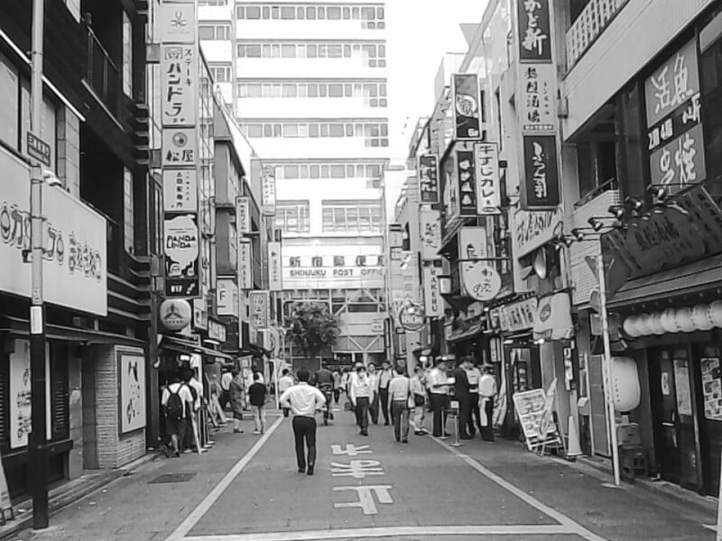 Black and white photo of a busy Japanese street lined with buildings and signs in Japanese; people in business attire walk and stand along the street, with a large building marked "SHINJUKU POST OFFICE" in the background.