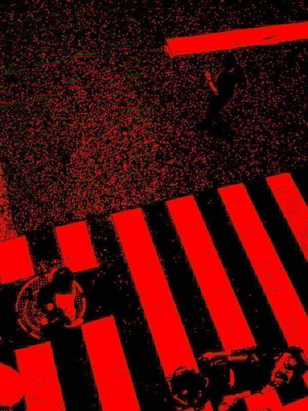 A high-contrast, red-and-black aerial photo shows people crossing a street at a crosswalk, with two distinct groups visible against the bold zebra stripes and dark asphalt.