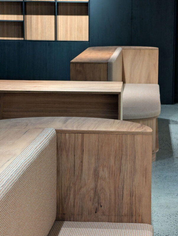 Banco moderno de madera con asientos tapizados de color beige ligeros, dispuestos en un interior simple, piso de concreto y estantes de madera en paredes oscuras en el fondo.