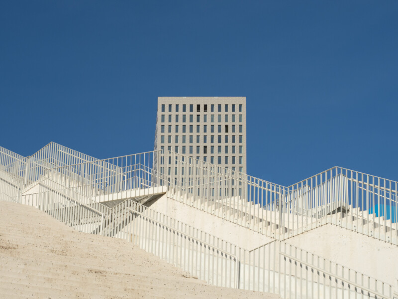 Geometric white stairs with metal railings lead upward, intersecting at angles, with a modern, grid-patterned building centered against a clear blue sky in the background.