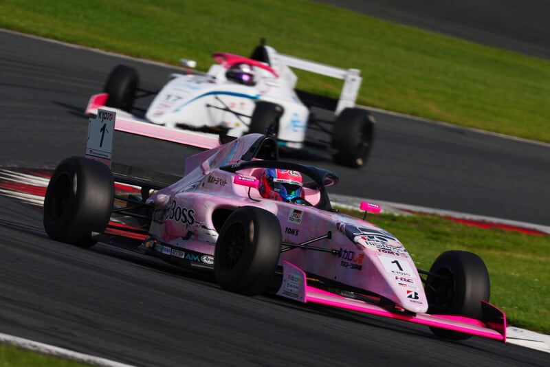 A pink and black formula race car leads on a racetrack, closely followed by a white car. Both drivers are wearing helmets, and the scene captures a high-speed moment with blurred grass and track in the background.