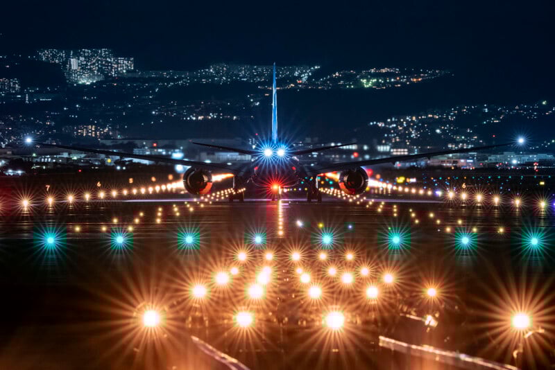 A commercial airplane is seen from behind, preparing for takeoff at night, with bright runway lights and city buildings glowing in the background.