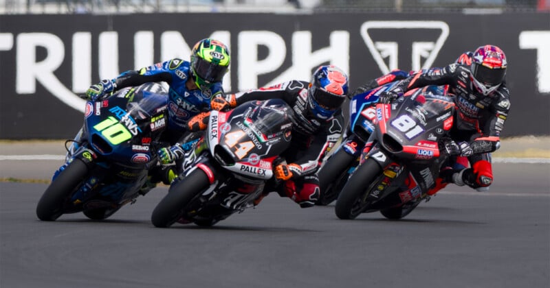 Three motorcycle racers in colorful racing suits and helmets lean sharply into a turn on a racetrack, competing closely. The background features a blurred "TRIUMPH" banner.