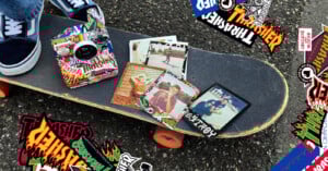 A skateboard with orange wheels rests on asphalt, covered in colorful "Thrasher" stickers. Several retro-style photos and more stickers are scattered on the skateboard. A person’s feet in blue sneakers are visible nearby.