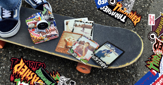 A skateboard with orange wheels rests on asphalt, covered in colorful "Thrasher" stickers. Several retro-style photos and more stickers are scattered on the skateboard. A person’s feet in blue sneakers are visible nearby.