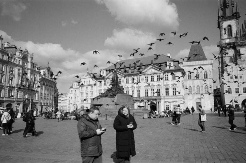 A city square with historic buildings, people walking and standing, and a flock of birds flying overhead. Two people in the foreground wear coats and appear to be checking their phones. The sky is partly cloudy.