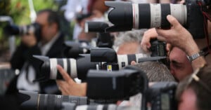 A group of photographers closely packed together, aiming large cameras with telephoto lenses, capturing images at an outdoor event.