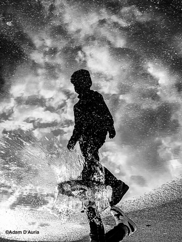La foto en blanco y negro muestra un reflejo de una persona que pasa a través de un charco, salpicando agua. El cielo nublado de arriba también se refleja en el charco, formando una escena surrealista y soñadora.