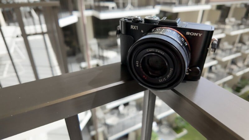 A Sony RX1 camera with a Carl Zeiss lens is placed on a metal railing, with an out-of-focus cityscape or building in the background.