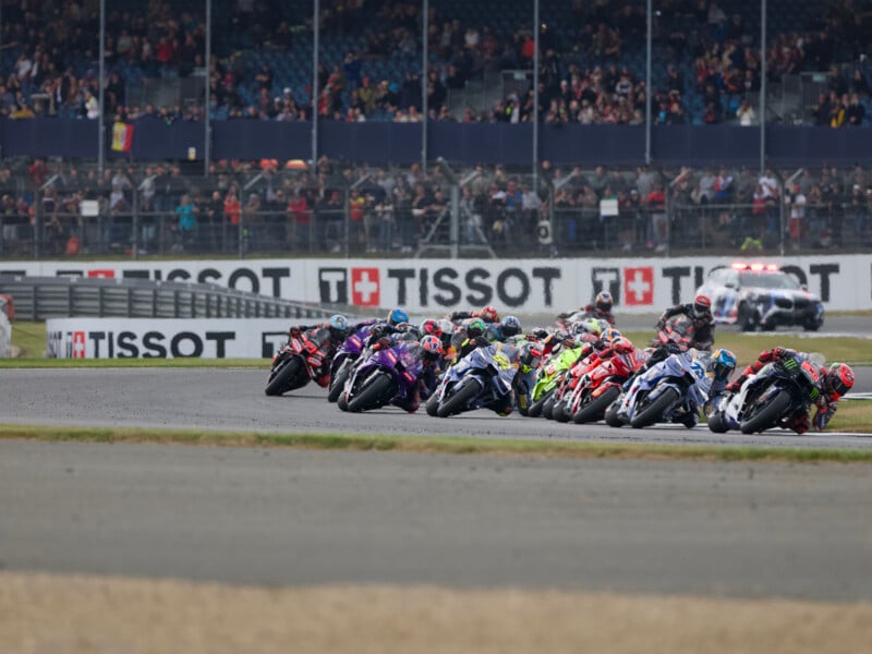 A group of motorcycle racers lean into a curve on a racetrack, closely packed together, with spectators watching from the stands in the background and Tissot advertisements visible along the barriers.