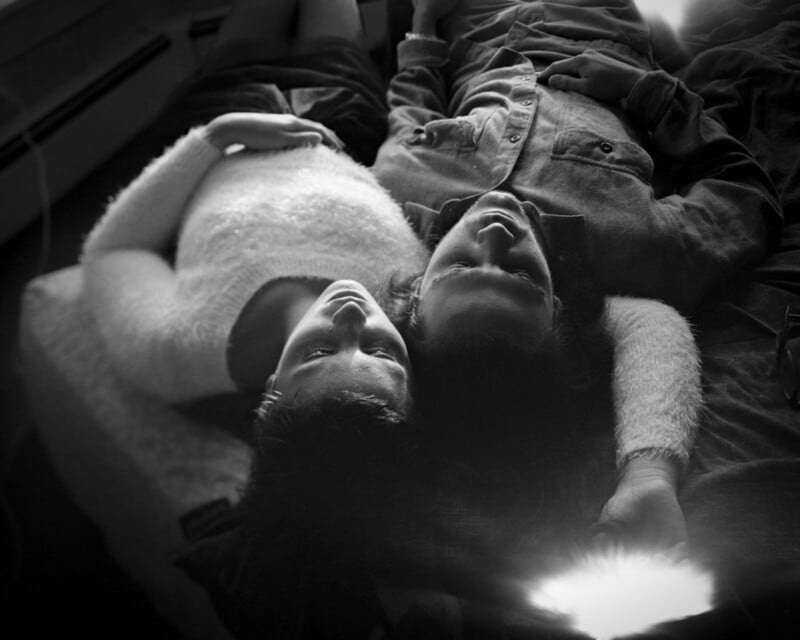 A black and white photo of two people lying close together on a bed, heads touching, looking relaxed and peaceful. One person wears a fuzzy sweater, the other a button-up shirt. Light flares softly in the image.