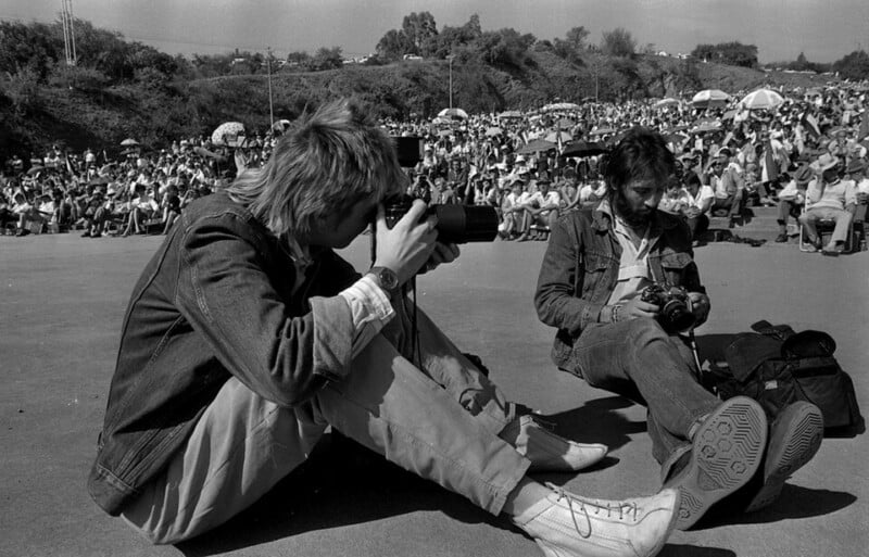Two photographers sit on the ground with cameras, focusing on the scene. A large crowd is gathered in the background at an outdoor event under sunny weather, with many people seated on grassy slopes.