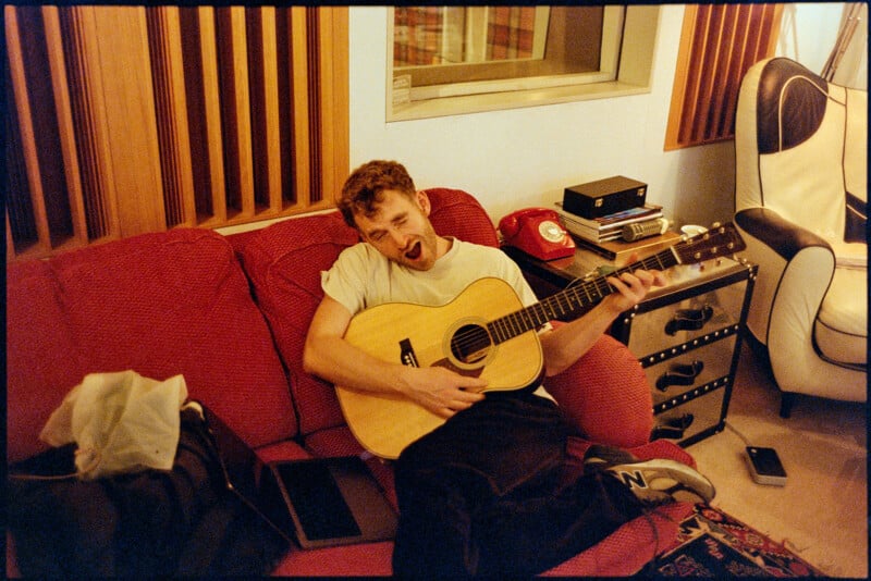Un hombre sentado en un sofá rojo tocando una guitarra y haciendo una cara divertida. A su alrededor hay una computadora portátil, teléfono y una caja de tejido. La habitación tiene paneles de madera, sillas blancas y un teléfono celular giratorio rojo.