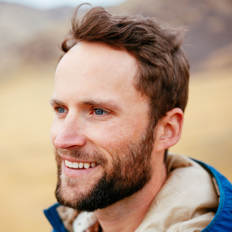 Un hombre con cabello marrón corto, sonriendo mientras mira a un lado. Está al aire libre con un abrigo azul con fondos borrosos, colinas y tonos de tierra.
