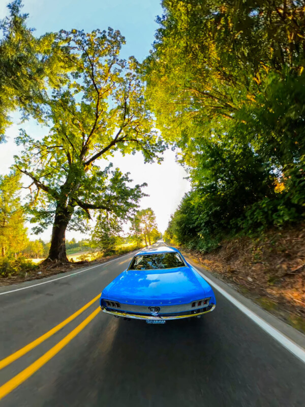 Un auto clásico azul brillante conduce a lo largo de una carretera de dos carriles rodeada de árboles verdes altos y hojas exuberantes, y el sol brilla de las hojas en un día despejado.