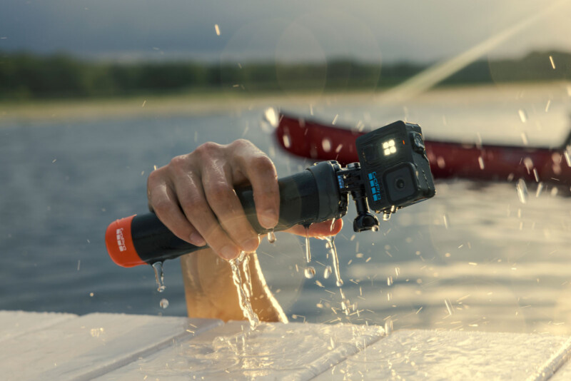 Una cámara de acción impermeable que sostiene una manija flotante montada sobre un mango flotante, goteando agua sobre el muelle, con una canoa roja borrosa y agua en el fondo.