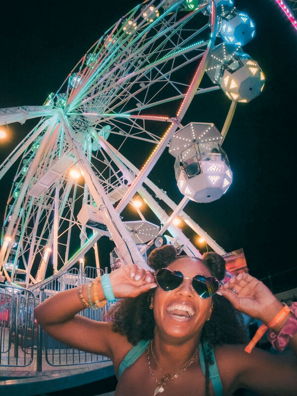 Una mujer con cabello rizado y gafas de sol en forma de corazón sonríe por la noche frente a la rueda de la fortuna brillantemente iluminada, creando un ambiente de carnaval divertido y festivo.