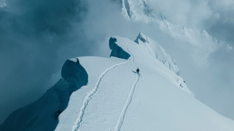 A lone climber walks along a narrow, snow-covered mountain ridge with deep footprints, surrounded by steep slopes and clouds in the background.