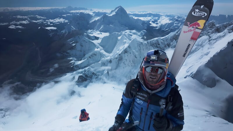 A climber in winter gear and sunglasses stands on a snowy mountain slope, carrying skis on his back. Another climber ascends below him, with rugged, snow-covered peaks and a dramatic sky in the background.