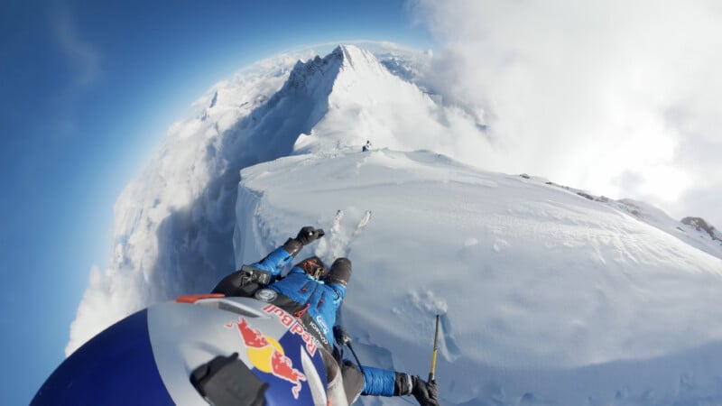 A mountaineer wearing blue gear and a helmet with a Red Bull logo climbs a snowy, narrow ridge high above the clouds, with dramatic mountain peaks and a curved horizon in the background.