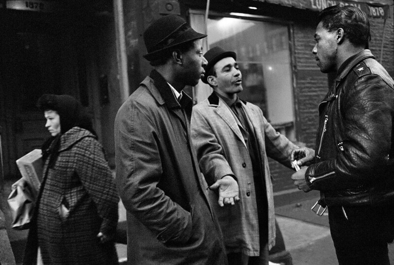 Three men stand talking on a city sidewalk; two wear coats and hats, one wears a leather jacket. A woman in a checked coat holding newspapers walks past in the background. The scene appears candid and urban.