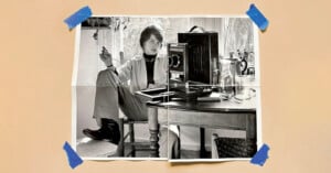 A black-and-white photo of a person sitting casually at a table with a camera and glass pitcher, taped at the corners to a beige surface with blue tape. The person is holding a cigarette and looking at the camera.