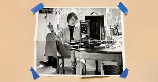 A black-and-white photo of a person sitting casually at a table with a camera and glass pitcher, taped at the corners to a beige surface with blue tape. The person is holding a cigarette and looking at the camera.