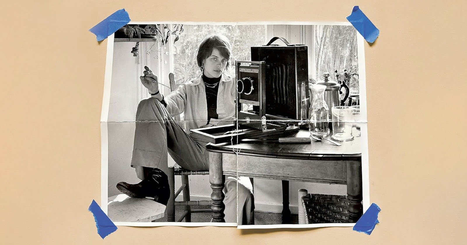 A black-and-white photo of a person sitting casually at a table with a camera and glass pitcher, taped at the corners to a beige surface with blue tape. The person is holding a cigarette and looking at the camera.