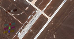 Satellite image of a desert airport runway and taxiway with aircraft lined up. Colorful rectangular markings are visible on the ground to the left of the runway complex.