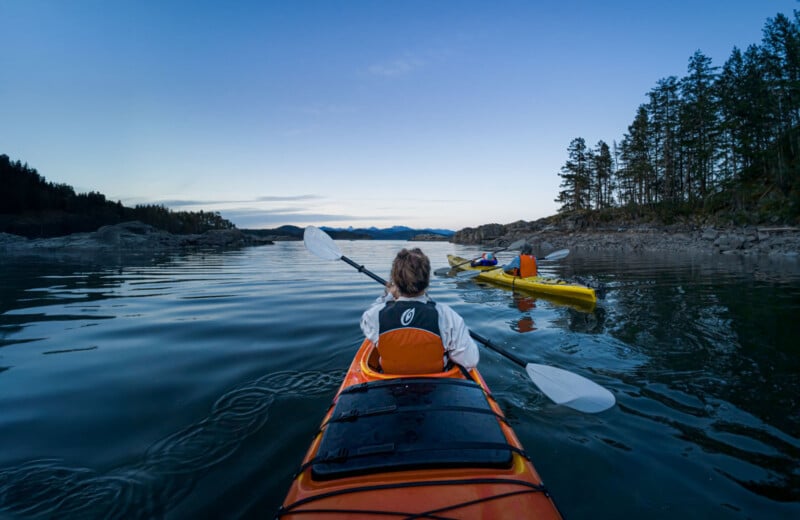 A person in an orange kayak paddles on calm water at dusk, following another kayaker in a yellow kayak. Pine trees and rocky shorelines border the tranquil lake under a clear sky.